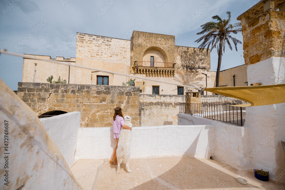 Fototapeta premium A woman and her white dog share a quiet morning moment on a sunlit rooftop, overlooking historic stone buildings and a palm tree in southern Italy