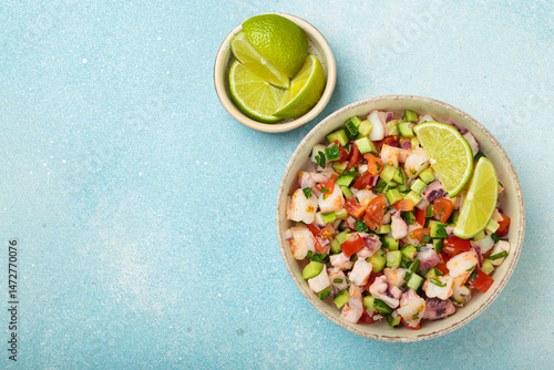 Fresh and vibrant peruvian seafood ceviche with shrimps, octopus, tomatoes, cucumber and parsley, served with lime in a bowl on a light blue background from above, dish of Peru. Copy space