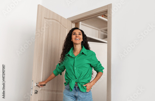Portrait of excited emotional lady walking in her apartment, entering new home, holding door knob. Happy young lady standing in doorway of modern flat, looking around at design interior, coming inside