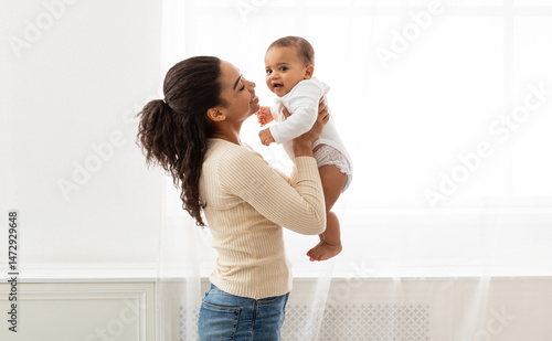 Mom's Love. Young Black Mother Holding Baby Toddler Playing And Cuddling With Little Infant Son Standing Near Window Indoor. Motherhood Happiness And Child Care, Maternity Leave Lifestyle. Side View