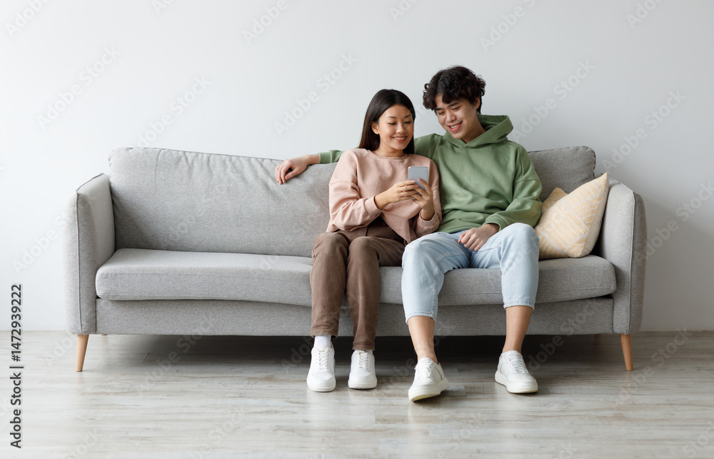 Happy young Asian couple using cellphone, browsing internet, chatting online, relaxing on sofa at home, free space. Cheery millennial family checking new mobile application, resting on couch together Happy young Asian couple using cellphone, browsing internet, chatting online, relaxing on sofa at home, free space. Cheery millennial family checking new mobile application, resting on couch together