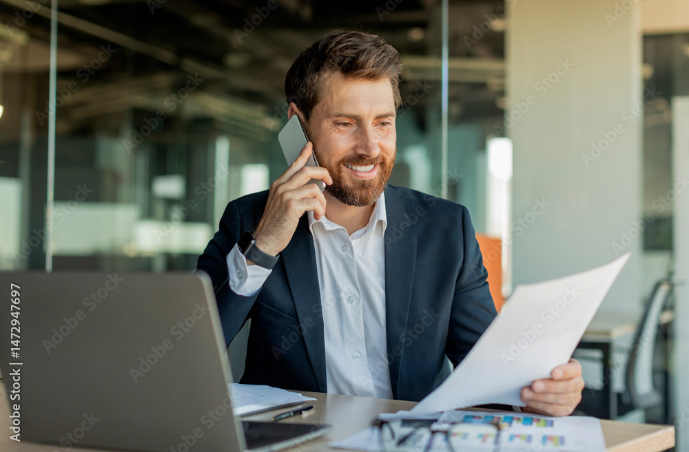 Corporate communication. Happy male manager working remotely, reading financial document and talking on mobile phone, sitting at desk in office interior Corporate communication. Happy male manager working remotely, reading financial document and talking on mobile phone, sitting at desk in office interior
