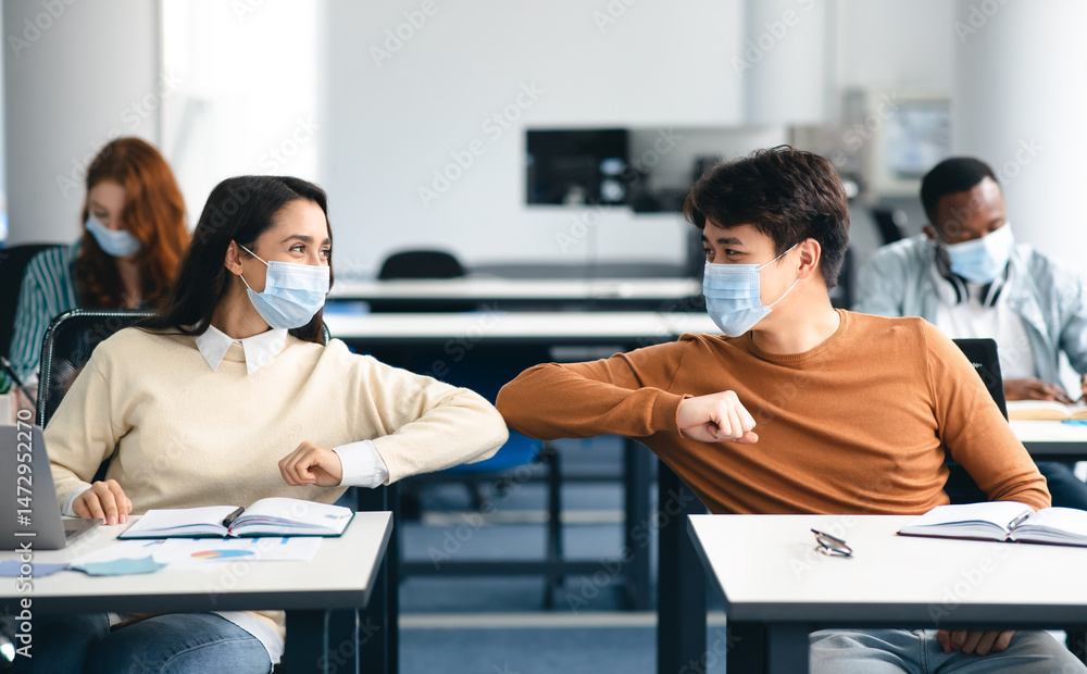Obraz premium Elbow Bump. Young multiethnic students wearing protective disposable surgical masks greeting each other at classroom. Man and woman avoid touching, studying in high school, sitting at table