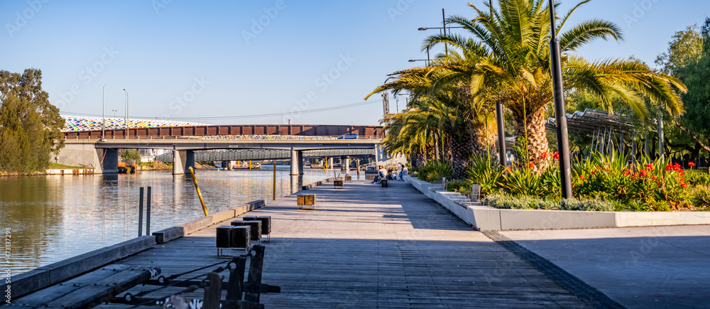 Fototapeta premium Footscray Wharf in Melbourne, Australia, with palm trees, landscaped gardens, and a pedestrian boardwalk beside the Maribyrnong River—riverside regeneration, public recreation, and modern urban design