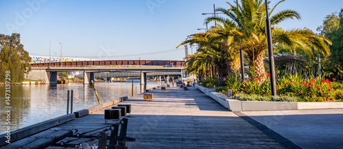 Fototapeta Naklejka Na Ścianę i Meble -  Footscray Wharf in Melbourne, Australia, with palm trees, landscaped gardens, and a pedestrian boardwalk beside the Maribyrnong River—riverside regeneration, public recreation, and modern urban design