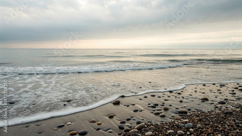 Serene coastal scene with gentle waves lapping against pebble strewn beach under cloudy sky, evoking calm and tranquility