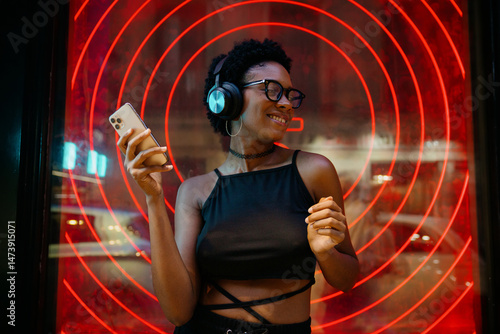 Young stylish Black woman vibing to playlist while streaming music, moving to rhythm with headphones on, neon-lit wall creating bold urban ambiance behind her