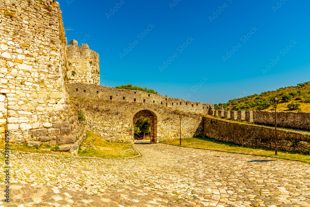 Obraz premium Fortified stone gateway of Berat Castle in Albania with cobblestone courtyard and ramparts atop a sunlit golden hillside under a clear azure sky