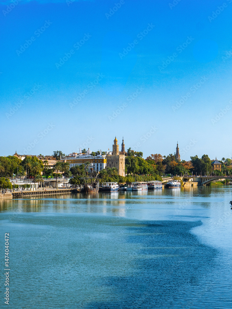 Fototapeta premium Scenic view of Canal de Alfonso XIII, Seville, Spain, with historic buildings and lush greenery reflecting on calm waters under a clear blue sky.