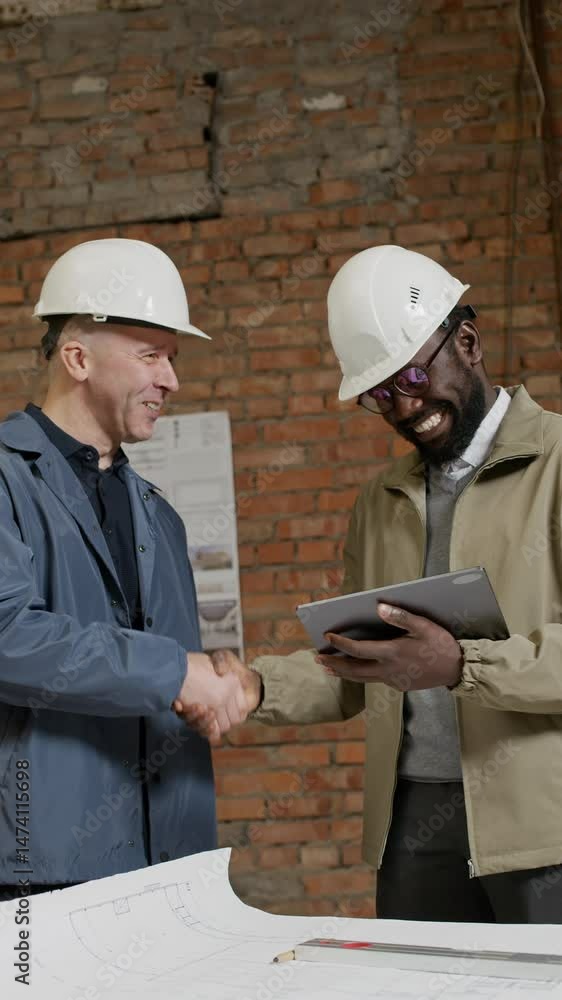 Vertical shot of diverse male architects in hardhats shaking hands using digital tablet when discussing blueprint on construction site Vertical shot of diverse male architects in hardhats shaking hands using digital tablet when discussing blueprint on construction site