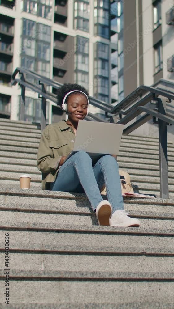 Smiling African American student in headset sitting on stairs with laptop during online class. Participating in video call or presenting project while sipping coffee and studying near university. Smiling African American student in headset sitting on stairs with laptop during online class. Participating in video call or presenting project while sipping coffee and studying near university.