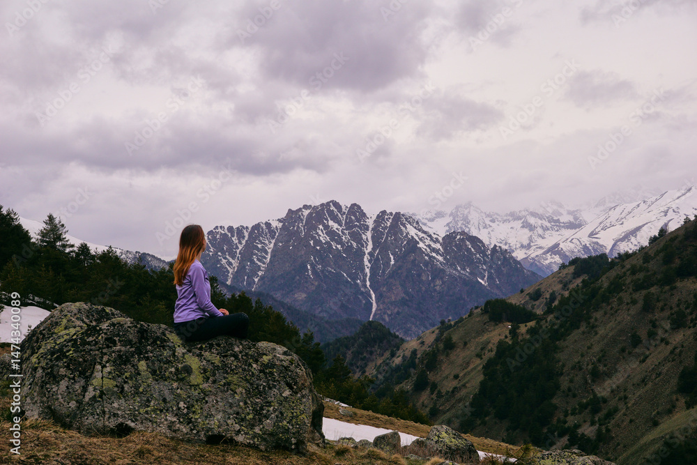 Naklejka premium A young woman sits on a large rock and looks at the mountains. Active travel in nature, hiking in the mountains