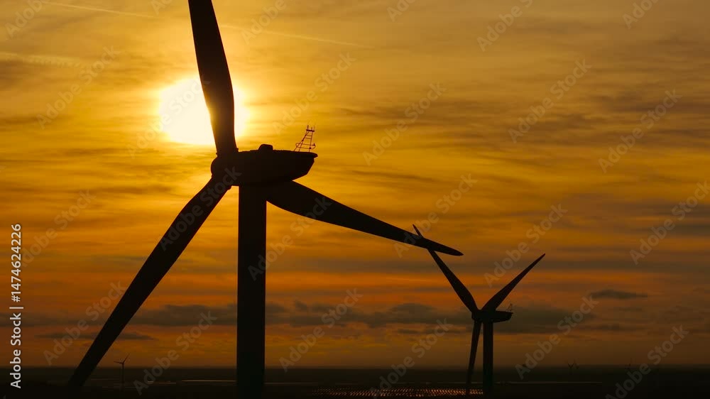 Wind farm generating clean renewable energy at sunset. Wind turbine turning circular blades to create green power under dramatic orange evening sky. Turning massive blades converting wind into