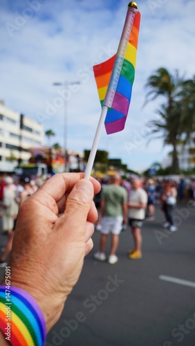 Hand holding small rainbow flag with rainbow wristband at Maspalomas Gay Pride with blurred crowd in background celebrating pride month