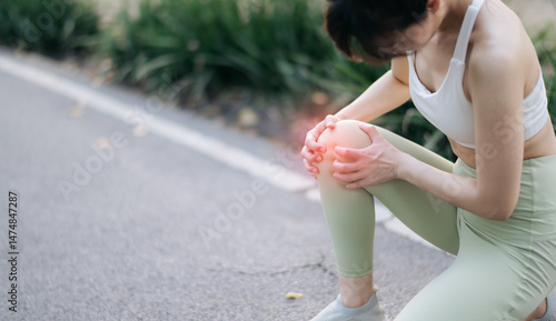 Asian woman experiencing knee pain during exercise, holding her knee with pained expression, wearing white sports bra and green leggings, on park pathway