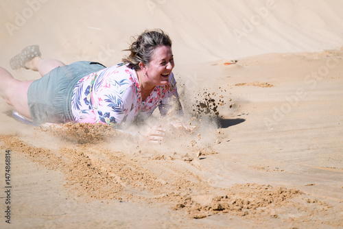 Middle age woman sandboarding down sand dune on boogie board, mid-life fun excitement, holiday vacation activity, healthy vital age ageing