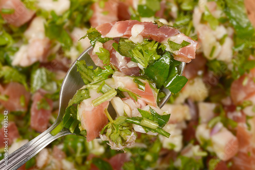 A spoonful of diced pancetta mixed with fresh green herbs and olive oil is held above a blurred background of the same mixture