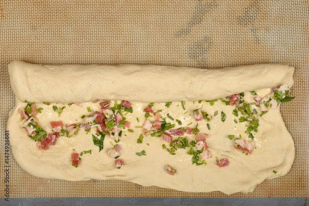 Raw dough is being rolled up with a filling of diced pancetta and green herbs on a baking mat, in the process of making pancetta rolls