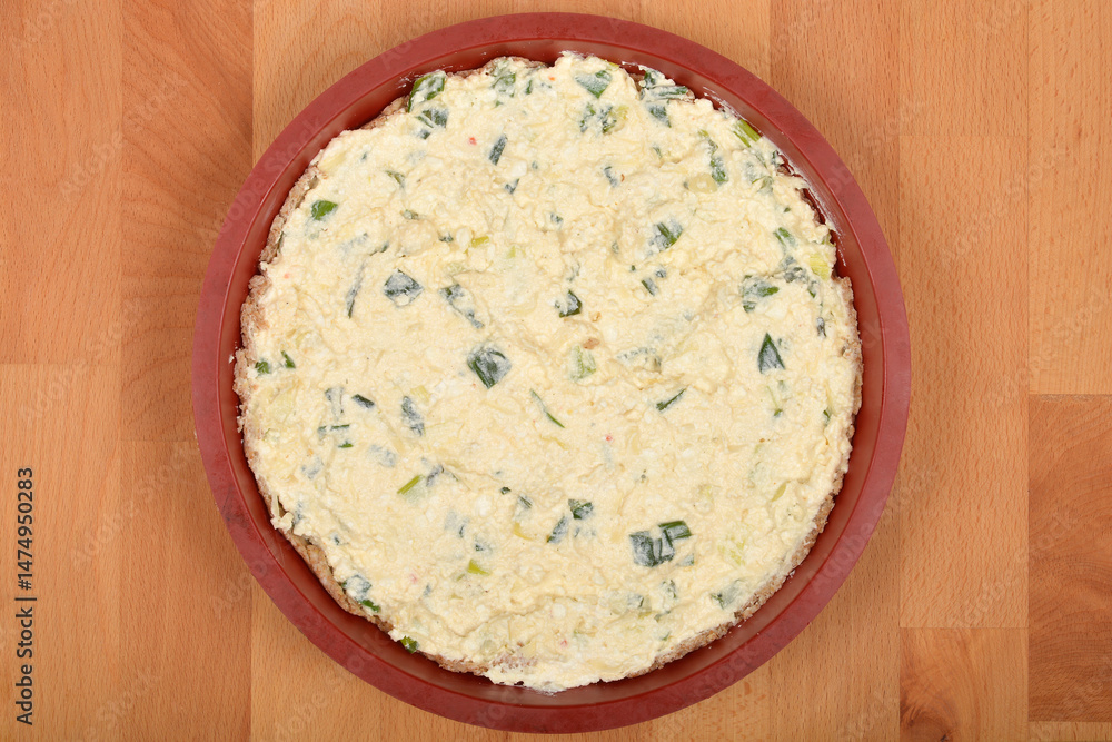 Raw cheese pie with green onionsA top-down view shows a round cheese pie with fresh cheese and green onions in a red baking dish on a wooden surface