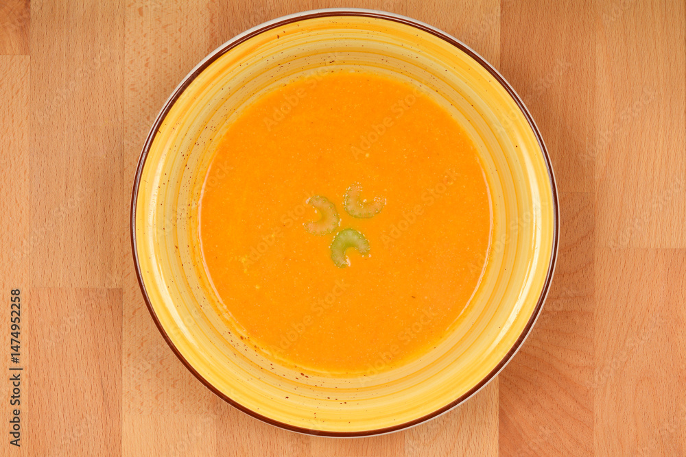A top-down view shows a creamy orange carrot soup garnished with celery in a yellow and brown bowl on a wooden surface