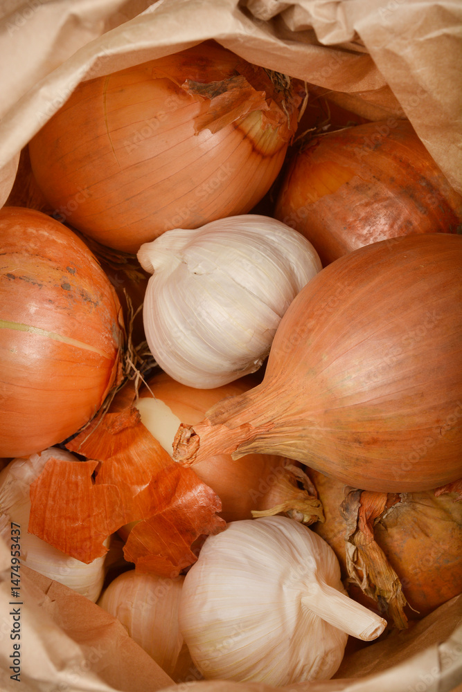 A close-up from inside a paper bag shows several brown onions and white garlic bulbs mixed together
