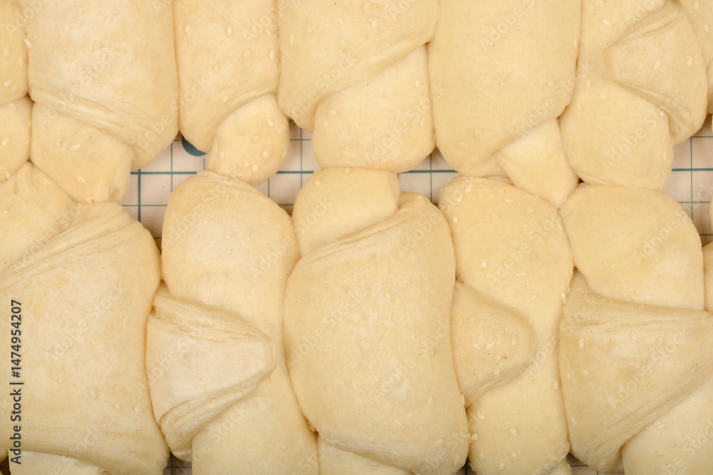 A close-up overhead shot shows rows of raw, pale yellow mini croissants sprinkled with sesame seeds on a grid surface