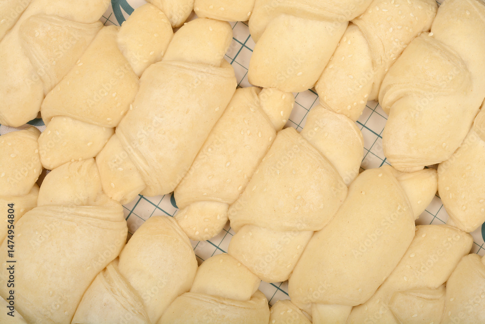 A close-up overhead shot shows rows of raw, pale yellow mini croissants sprinkled with sesame seeds on a grid surface