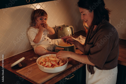 In a warm kitchen, a mother slices apples while her child sits on the counter, looking curious and playful. The setting is intimate and inviting, highlighting bonding moments