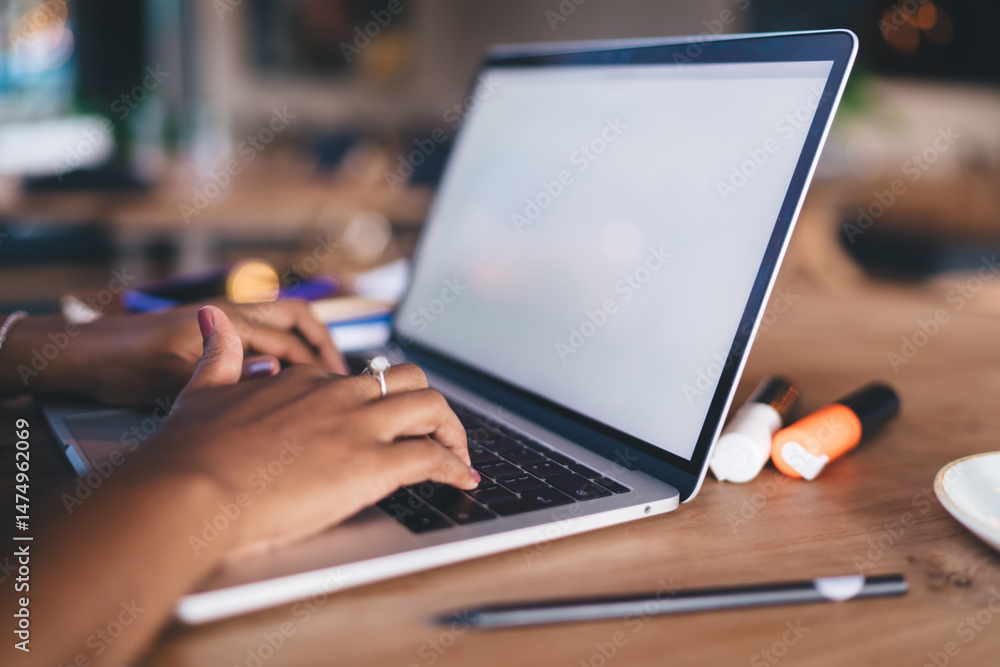 Close-up of hands typing on laptop keyboard during remote work session in cafe, surrounded by highlighters, documents, and a cup of coffee on wooden desk surface