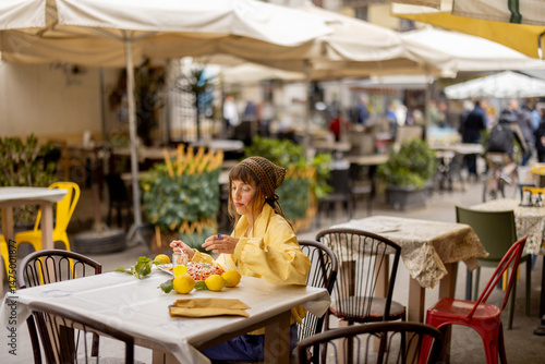 Quadro su tela A woman in a yellow jacket enjoys a plate of pasta alone at a street cafe under large umbrellas