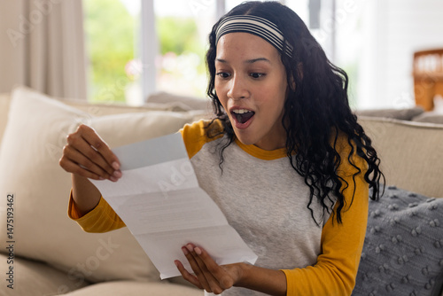 Excited woman reading letter on couch, expressing surprise and happiness at home