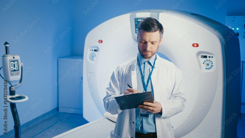 Positive Caucasian male in medical coat holding clipboard while posing near CT scanner. Preparing for next appointment or reviewing patient files inside clinical diagnostic center. Positive Caucasian male in medical coat holding clipboard while posing near CT scanner. Preparing for next appointment or reviewing patient files inside clinical diagnostic center.