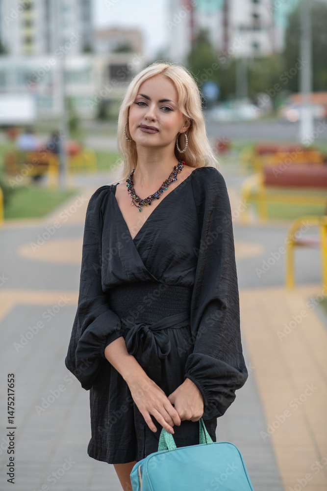Fototapeta premium Portrait of a young beautiful blonde girl in a dark dress in a city dress in the summer.