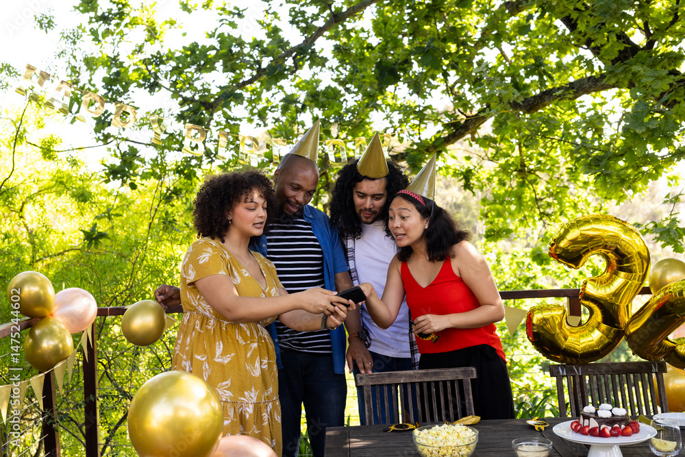 Fototapeta premium Friends celebrating birthday outdoors, wearing party hats and looking at smartphone
