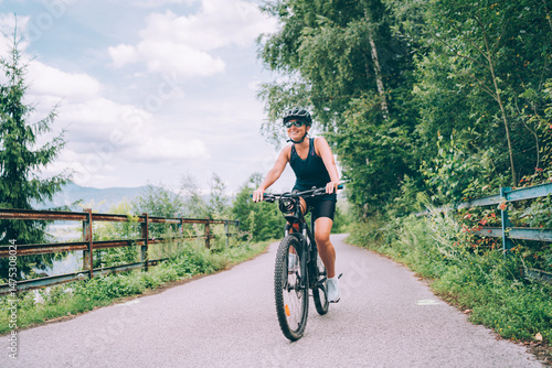 Fotografie Portrait of happy smiling woman dressed in cycling clothes, helmet and sunglasses riding bicycle on asphalt out-of-town bicycle path