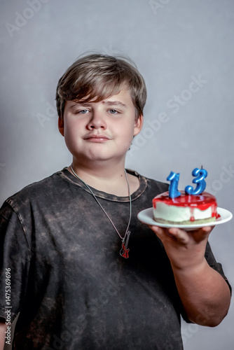 Young person holding small cake adorned with blue candles shaped as numbers 1 and 3, symbolizing age thirteen. Wearing dark T-shirt and pendant, expression is neutral and confident