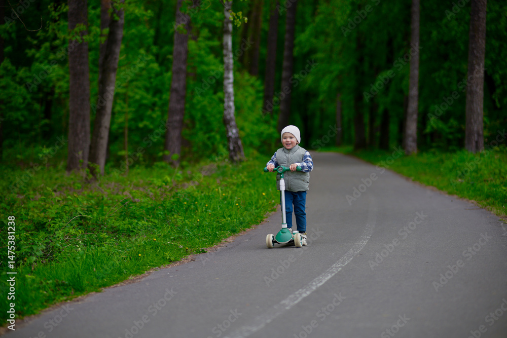 Obraz premium Little boy riding a green scooter on a paved path through a lush forest. Child dressed in a vest, beanie, and sneakers enjoying outdoor playtime in nature. Fun and active childhood moment.