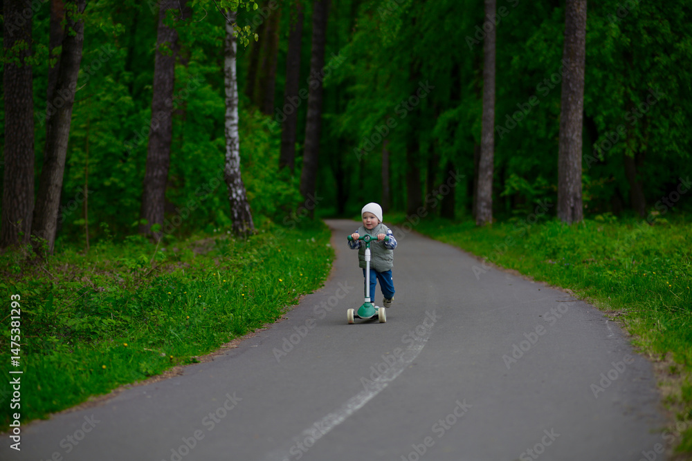 Fototapeta premium Little boy riding a green scooter on a paved path through a lush forest. Child dressed in a vest, beanie, and sneakers enjoying outdoor playtime in nature. Fun and active childhood moment.