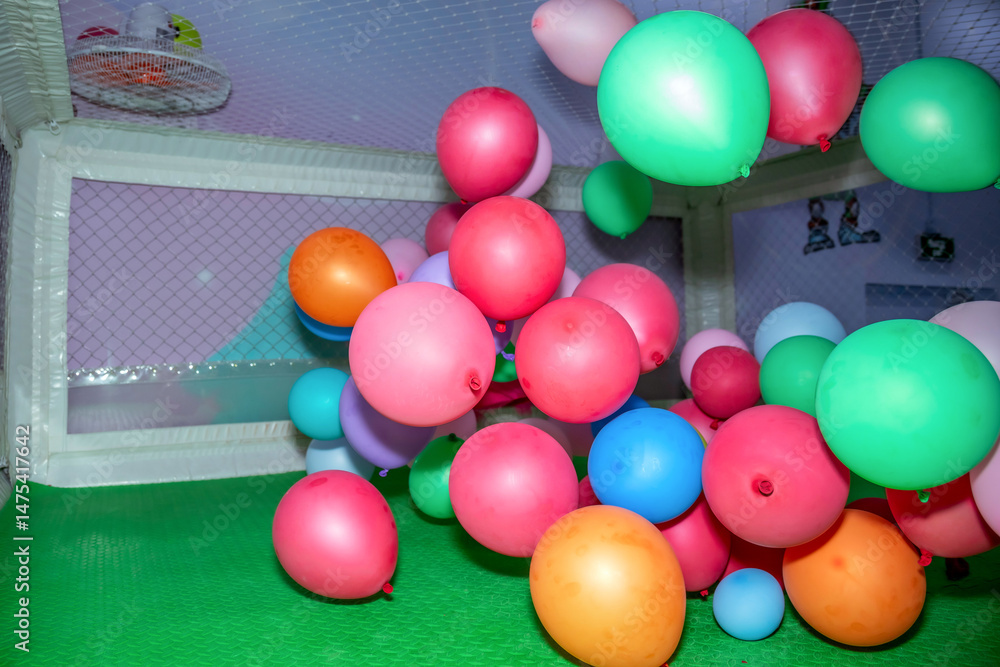 Obraz premium Colorful balloons of various sizes are scattered across a green play area, enclosed by a netted fence. Bright hues of pink, blue, green, and orange are prominently featured