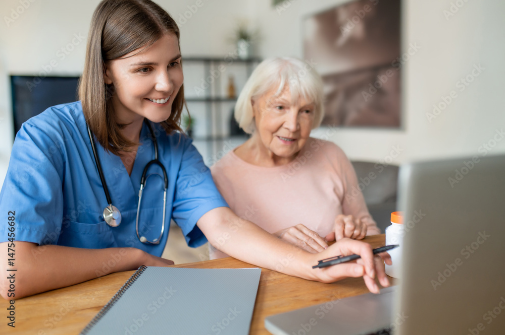 Fototapeta premium Female nurse in uniform using laptop with elderly patient in offering support for senior woman