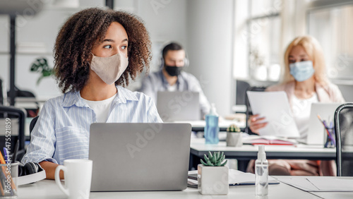 Creating an idea at work brainstorming for project. Serious african american lady in protective mask looks at window working at computer in modern office during outbreak of coronavirus, copy space