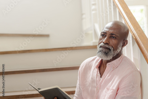 Elderly man sitting on stairs, reading book and reflecting peacefully at home