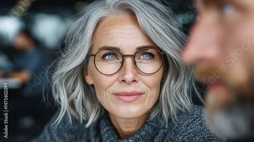 Thoughtful older woman with grey hair wearing glasses in a modern indoor setting, looking to the side with a contemplative expression, natural light ambiance