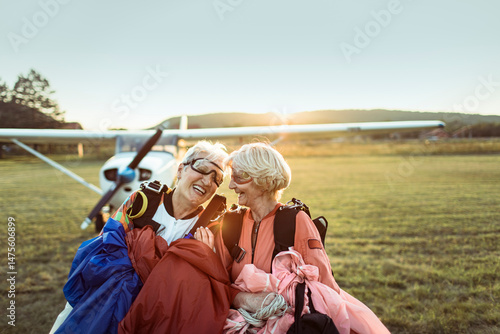 A senior lesbian couple stands on a field, airplane in the backdrop, beaming with pride and exhilaration after skydiving. Their smiles hint at a bucket list dream fulfilled.