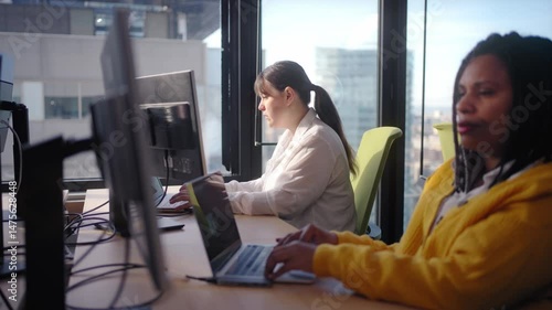 Two female programmers are working on their computers in a modern office with large windows, collaborating on a project and focused on their tasks