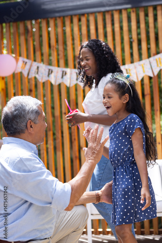 Grandfather playing with granddaughter, mother smiling at outdoor birthday party