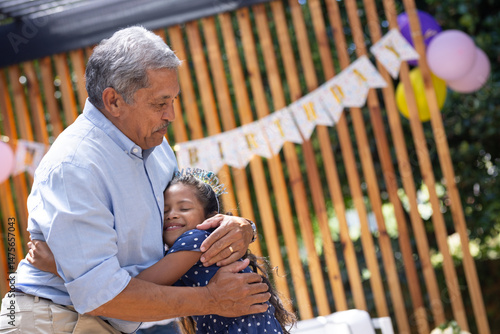 Grandfather hugging granddaughter at outdoor birthday party, both smiling warmly