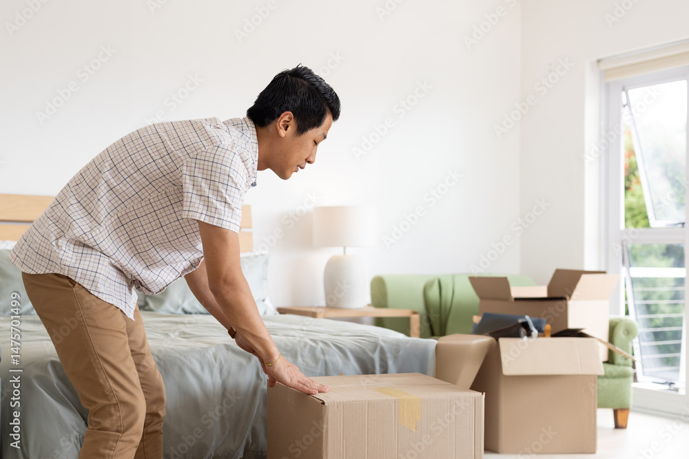 Mid adult Asian man bending over lifting cardboard box in bedroom with wooden headboard and lamp Mid adult Asian man bending over lifting cardboard box in bedroom with wooden headboard and lamp