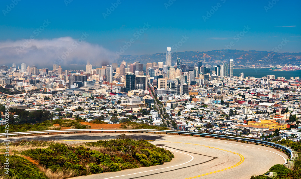 Obraz premium Panoramic view of downtown San Francisco from Twin Peaks, featuring iconic skyscrapers, urban density, coastal fog, and the curving summit roadway under a clear summer sky.