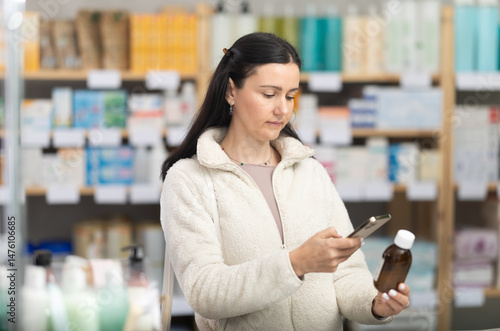 Adult woman in an autumn jacket scans cough syrup on her phone to read detailed instructions and ingredients on the pharmacy's website. Pharmacy visitor pays for purchases using a qr code